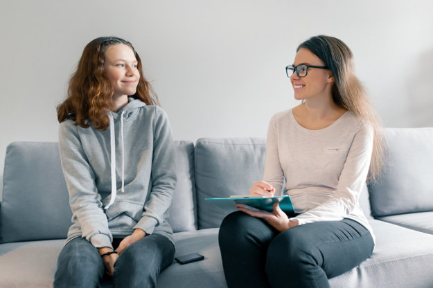 Young woman professional psychologist talking with teenager girl 14, 15 years old sitting in office on sofa. Mental health of child in adolescence young adult works with therapist at residential treatment center in california